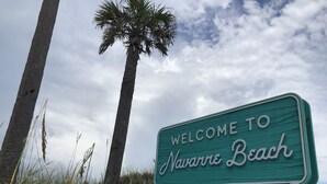 Un cartel de bienvenida a Navarre Beach, Florida, con palmeras y césped al fondo.