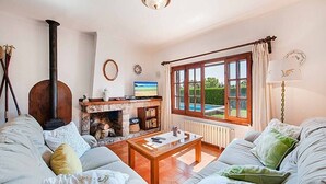 A cozy living room with a fireplace, a sofa set, a coffee table, and a view of a garden through a large window. - Villa Provenza, Cozy villa with pool near Port Pollensa (Port de Pollença)