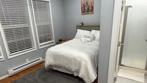 Ein Schlafzimmer mit Bett, grauen Wänden, weißen Jalousien, einem grauen Teppich und einer Tür.