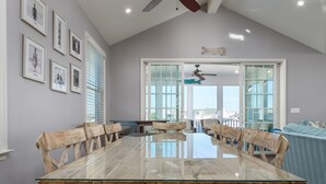 Un comedor amplio con una gran mesa de vidrio y sillas de madera, un ventilador de techo y vistas amplias a través de ventanas grandes.