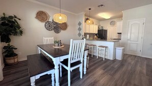 Un comedor con mesa de madera y sillas blancas, una cocina con armarios blancos y una puerta blanca.