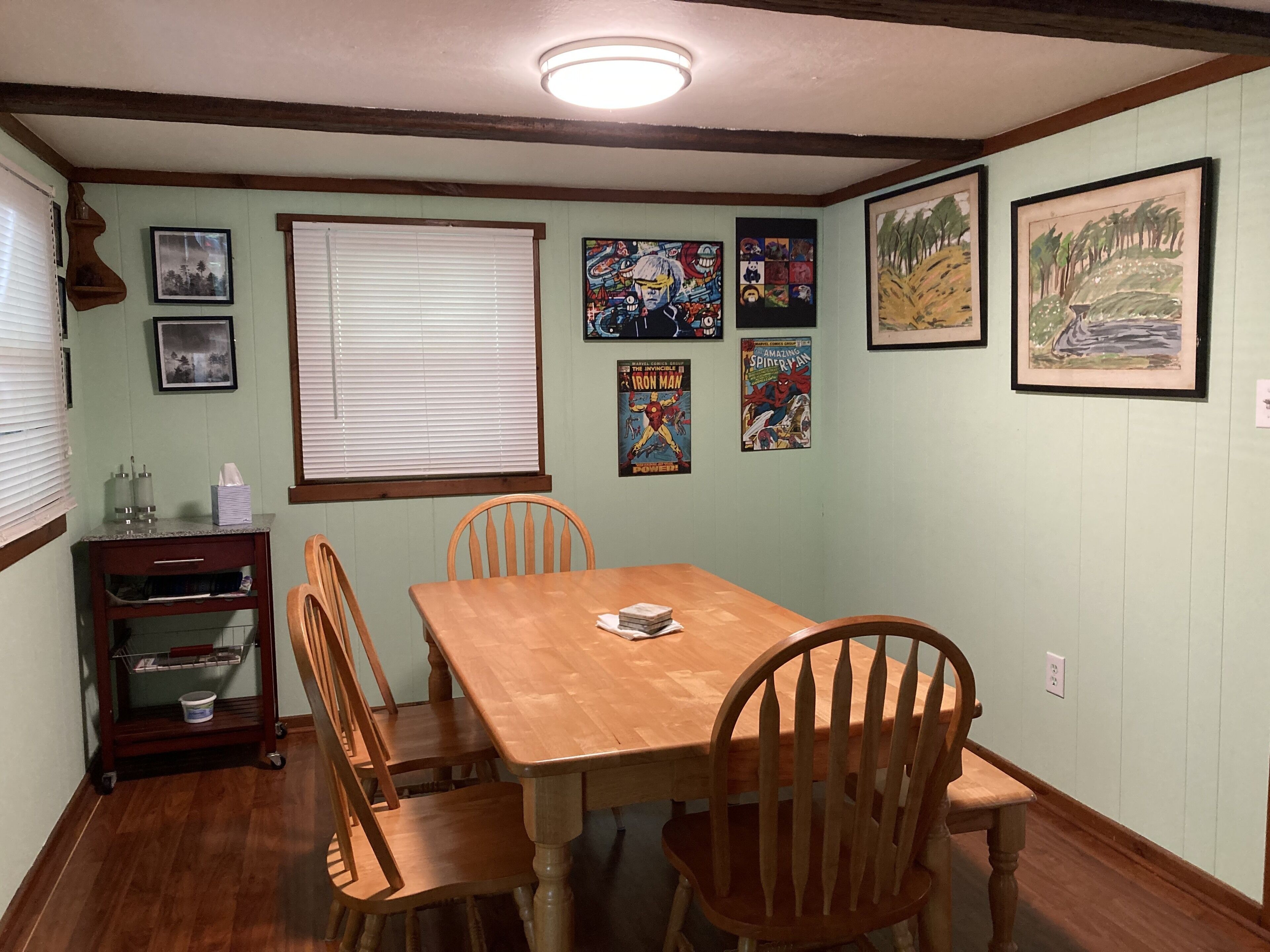 A dining room with a wooden table and chairs, a window with blinds, and various framed artworks on the walls.