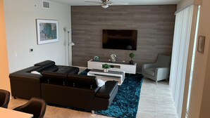 Ein modernes Wohnzimmer mit einem Ecksofa, einem Couchtisch, einem an einer Holzwand montierten Fernseher, einem Deckenventilator und einem großen Fenster mit Vorhängen.