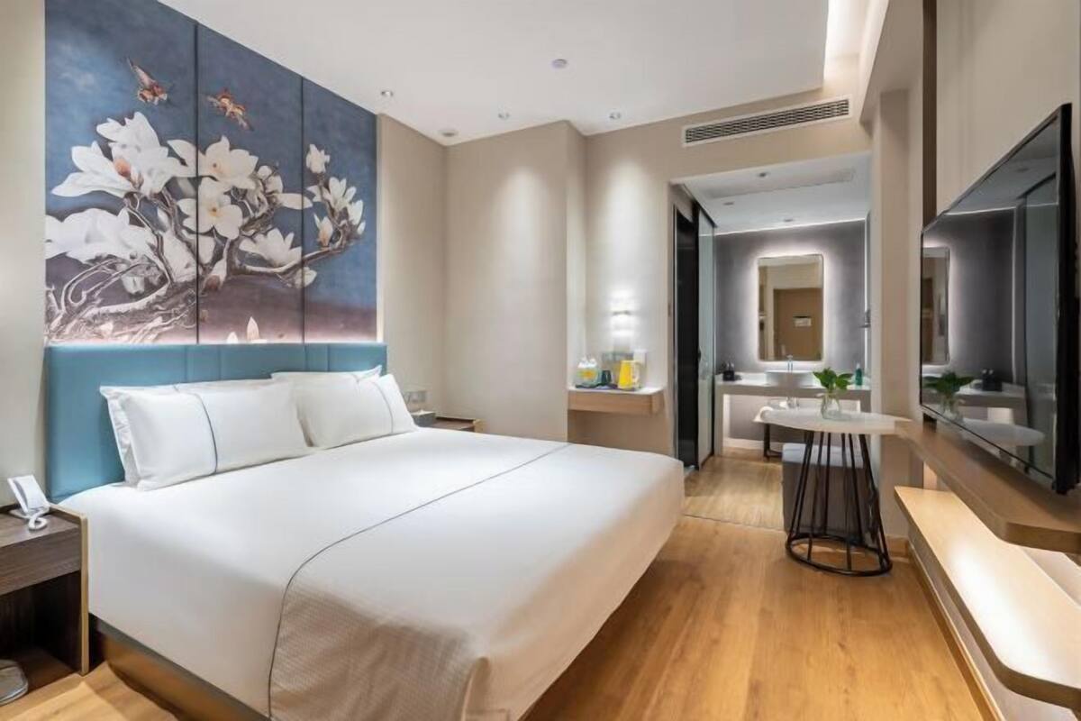 A modern hotel room with a large bed, a painting of flowers, a small table with a plant, and a bathroom visible through an open door.