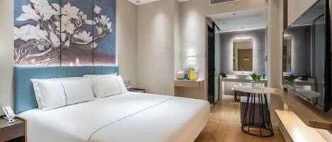 Habitación de hotel con una cama grande, un cuadro de flores, un escritorio, una silla y un baño con espejo y lavamanos.