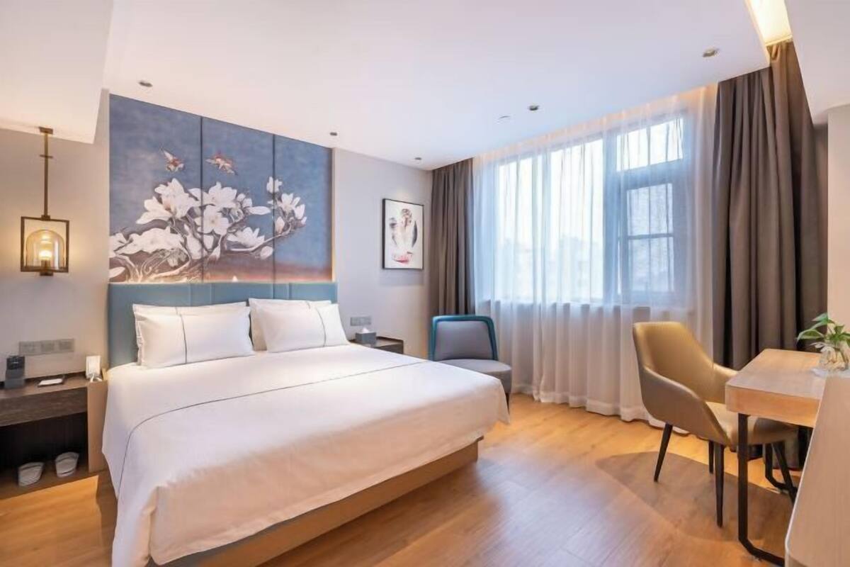 A modern hotel room with a large bed, a desk, and a chair. There is a painting on the wall and a window with curtains.