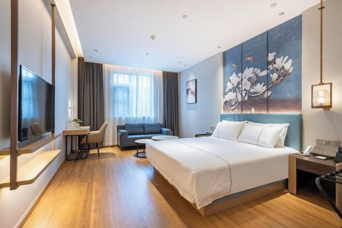 A modern hotel room with a large bed, a desk, a sofa, and a painting on the wall.