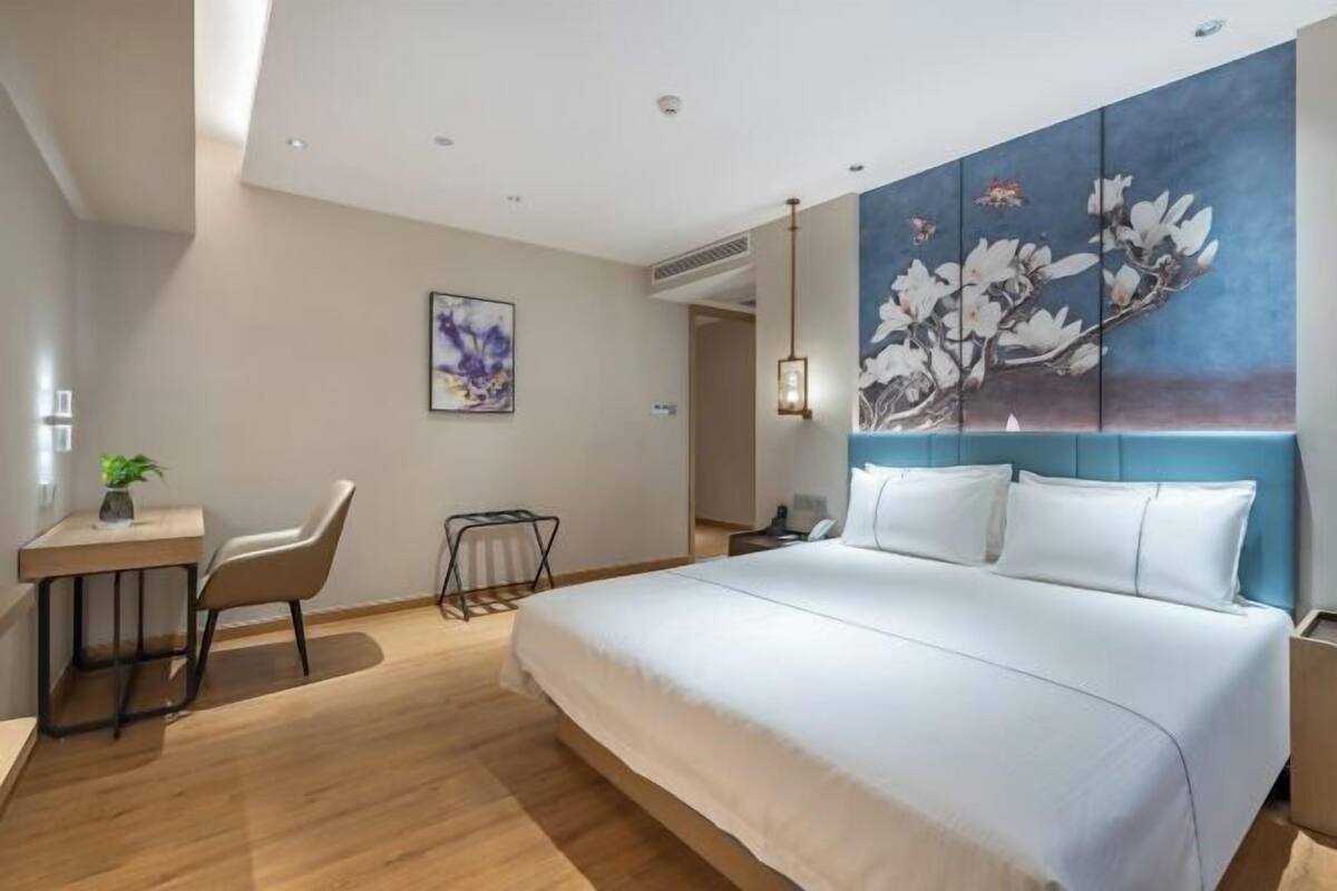 A hotel room with a large bed, a desk with a chair, a small table with a plant, and a painting on the wall.