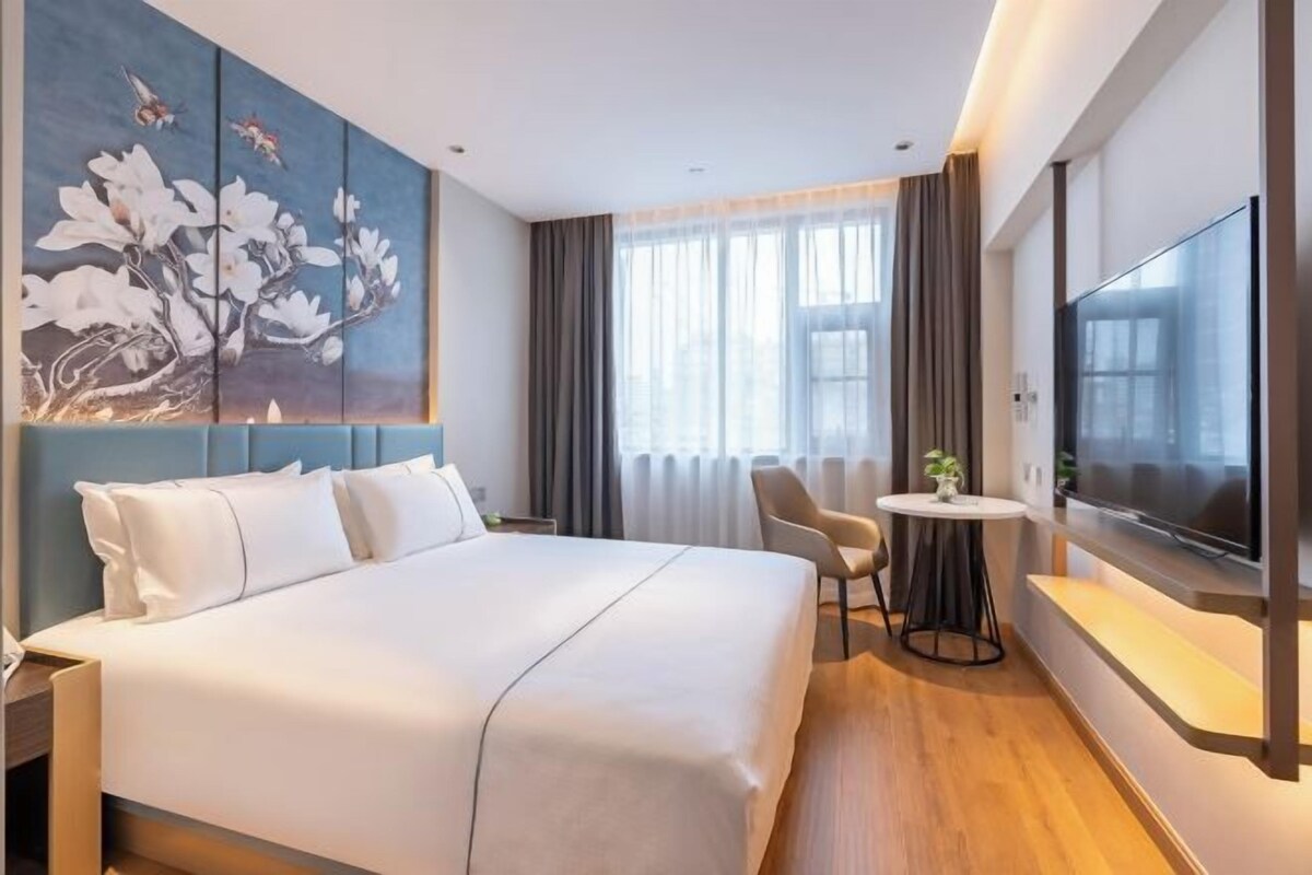 A modern hotel room with a large bed, a flat-screen TV, a small round table with a chair, and a painting on the wall.