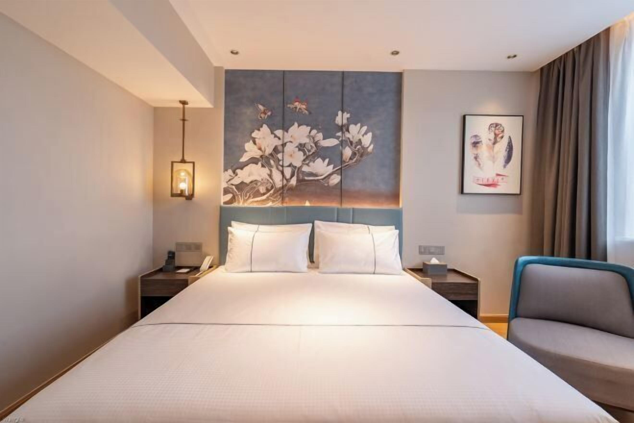 Quarto de hotel com uma cama grande, uma mesa de cabeceira com abajur, uma cadeira e um quadro na parede.