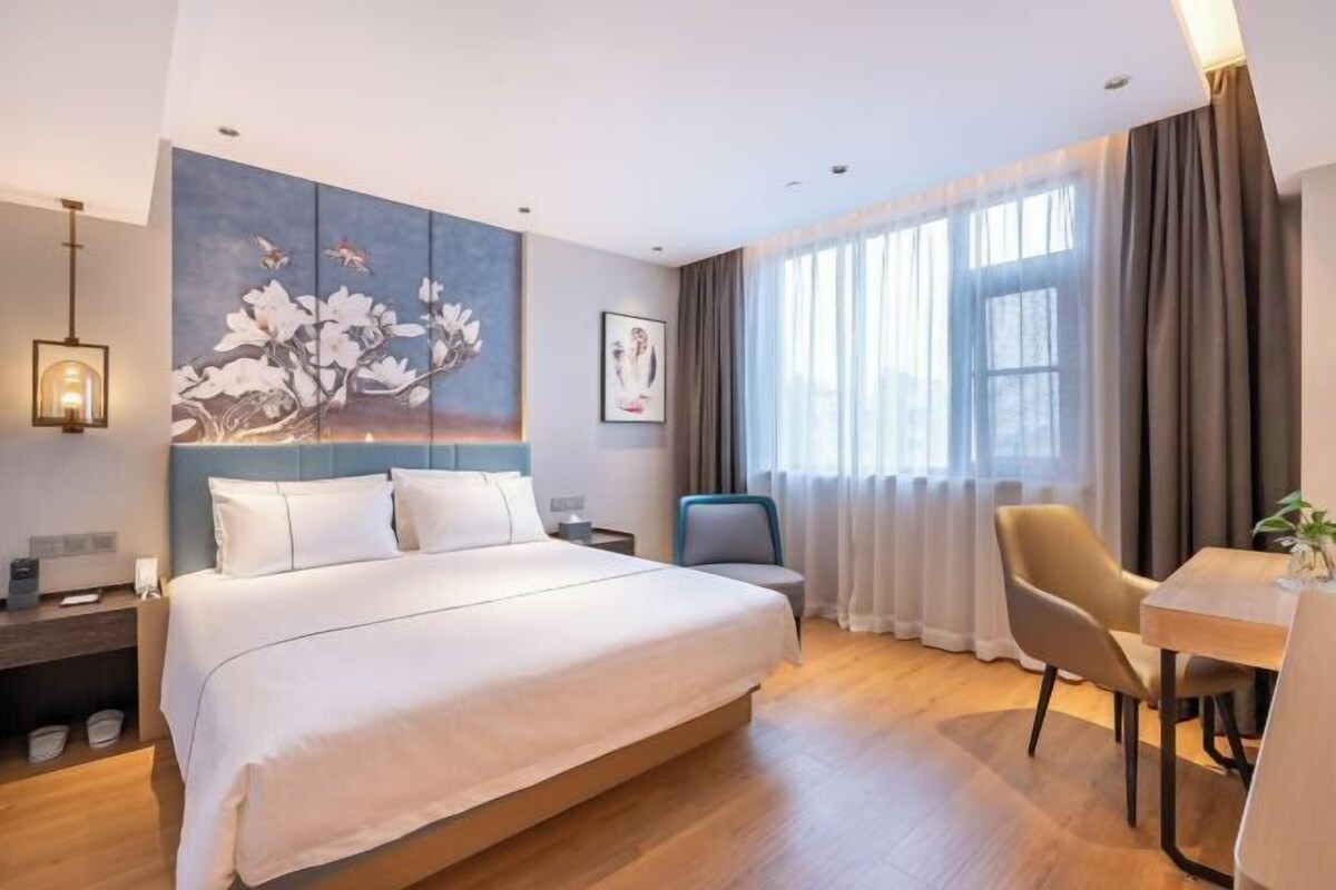 A modern hotel room with a large bed, a desk, and a chair. There is a painting on the wall and a window with curtains.