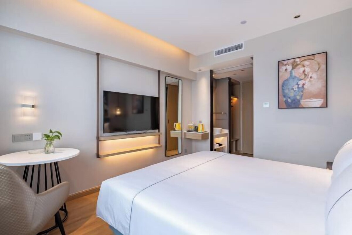 A modern hotel room with a large bed, a TV mounted on the wall, a small round table with a plant, and a painting on the wall.
