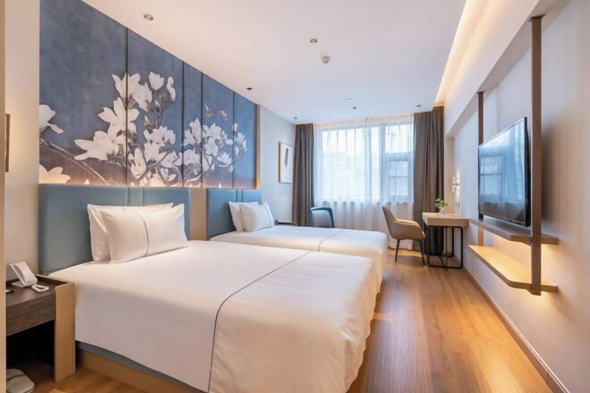 A hotel room with two beds, a desk, a chair, a TV, and a large floral artwork on the wall.