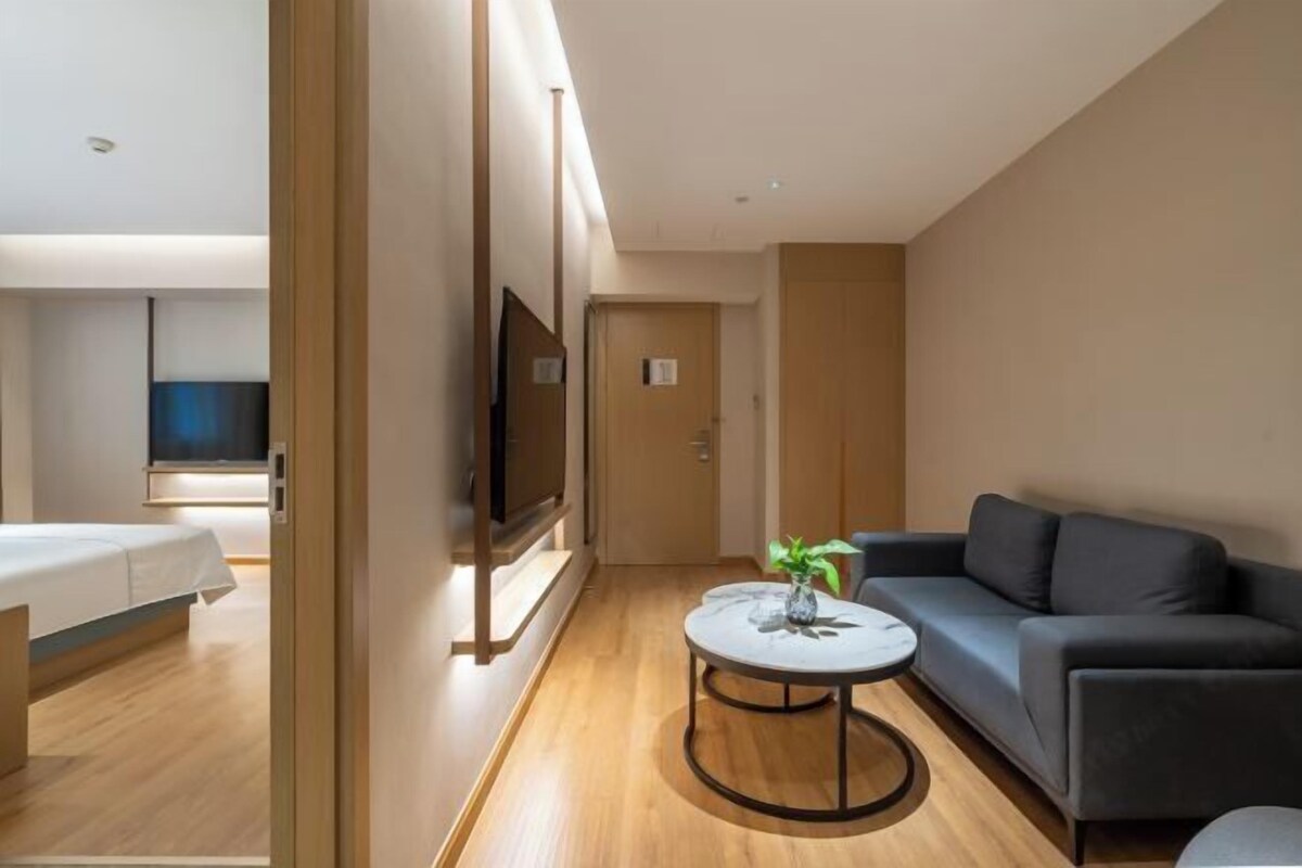 A modern hotel room with a wooden floor, a grey sofa, a small round table with a vase, and a TV mounted on the wall.