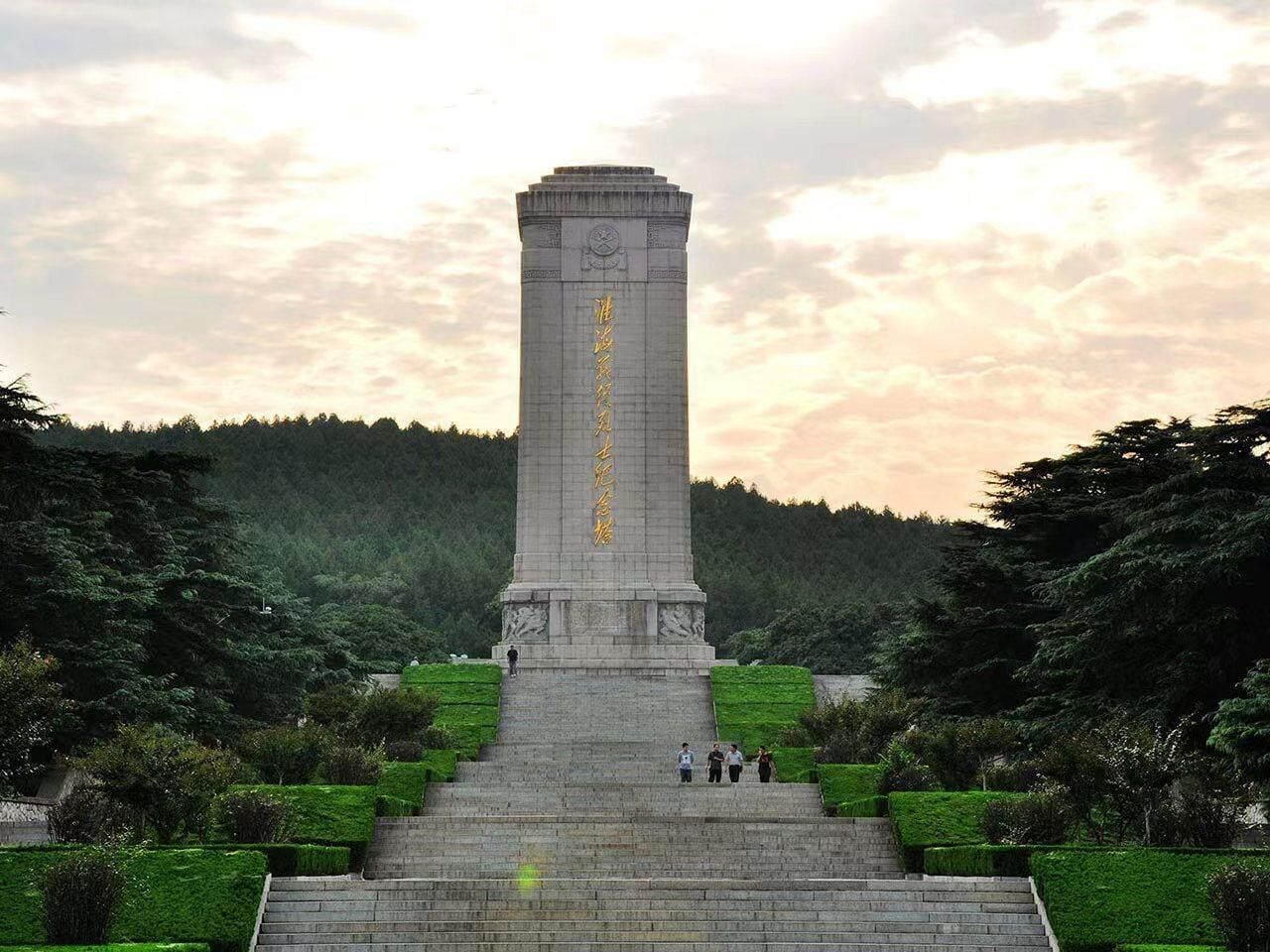 Un monumento alto con inscripciones, rodeado de un jardín cuidado con escaleras que conducen hasta él.