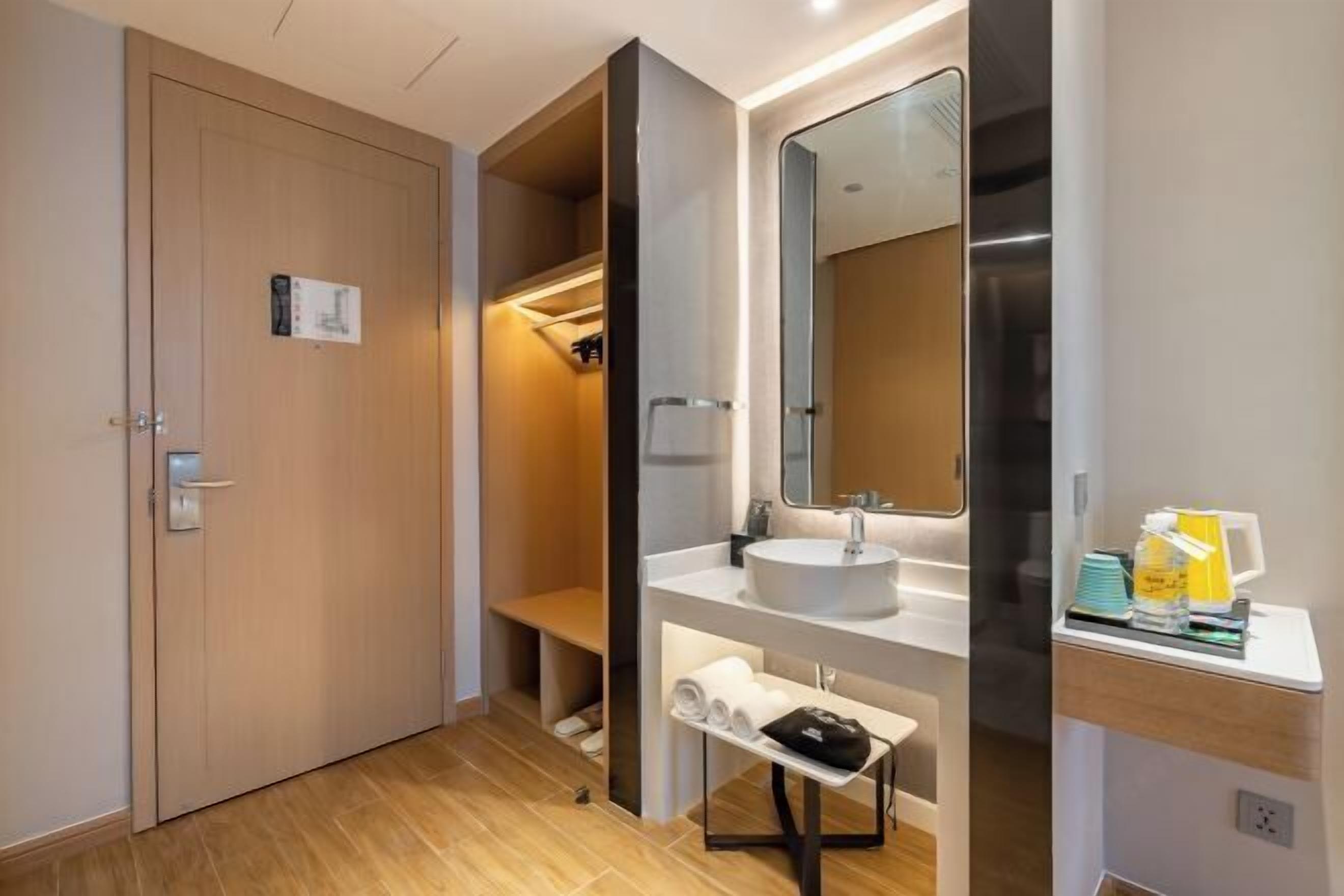 Een moderne badkamer met een houten deur, een bankje, een wastafel met spiegel en een plank met handdoeken en toiletartikelen.