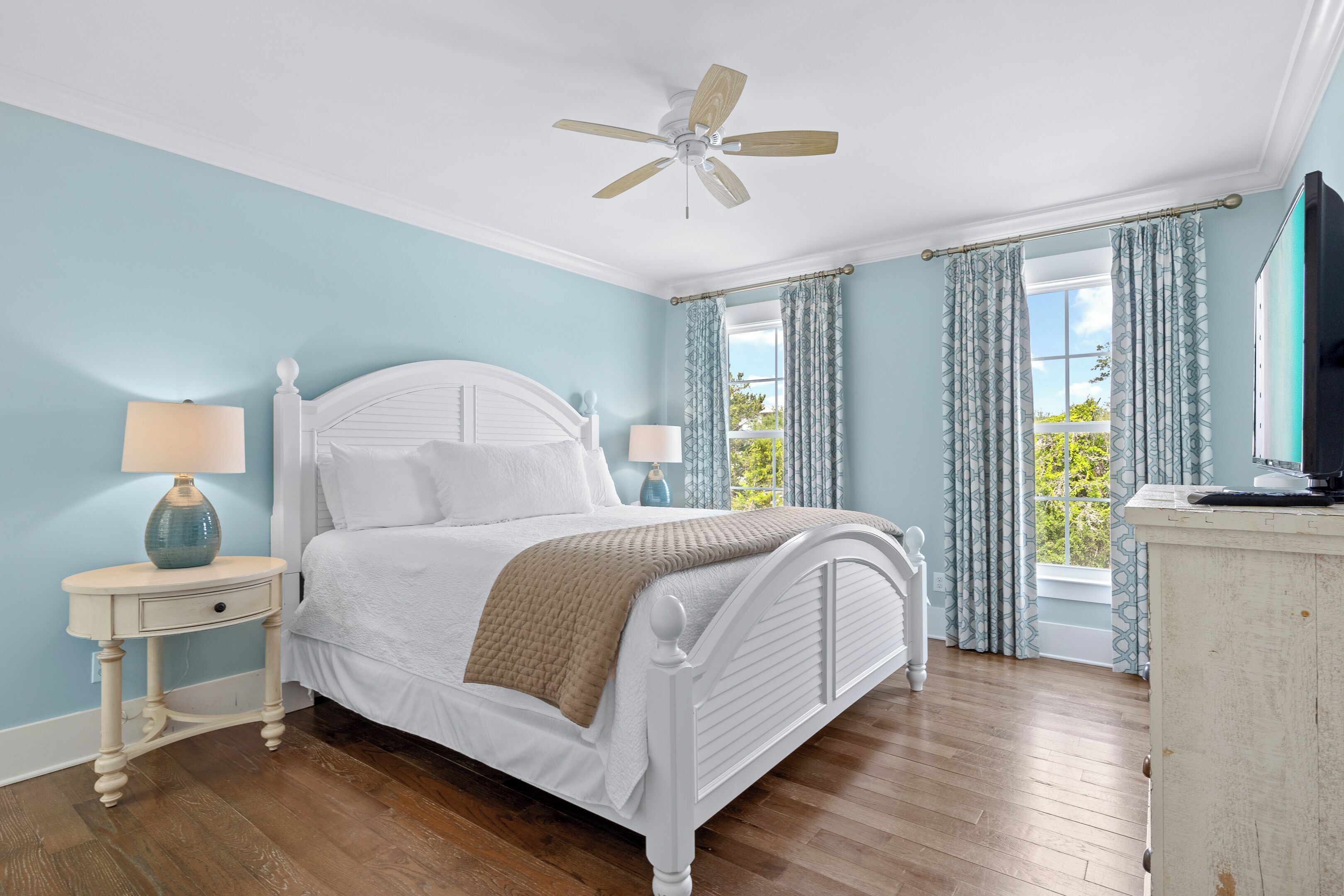 Une chambre à coucher avec un lit blanc, deux tables de chevet, un ventilateur de plafond et une fenêtre avec des rideaux.