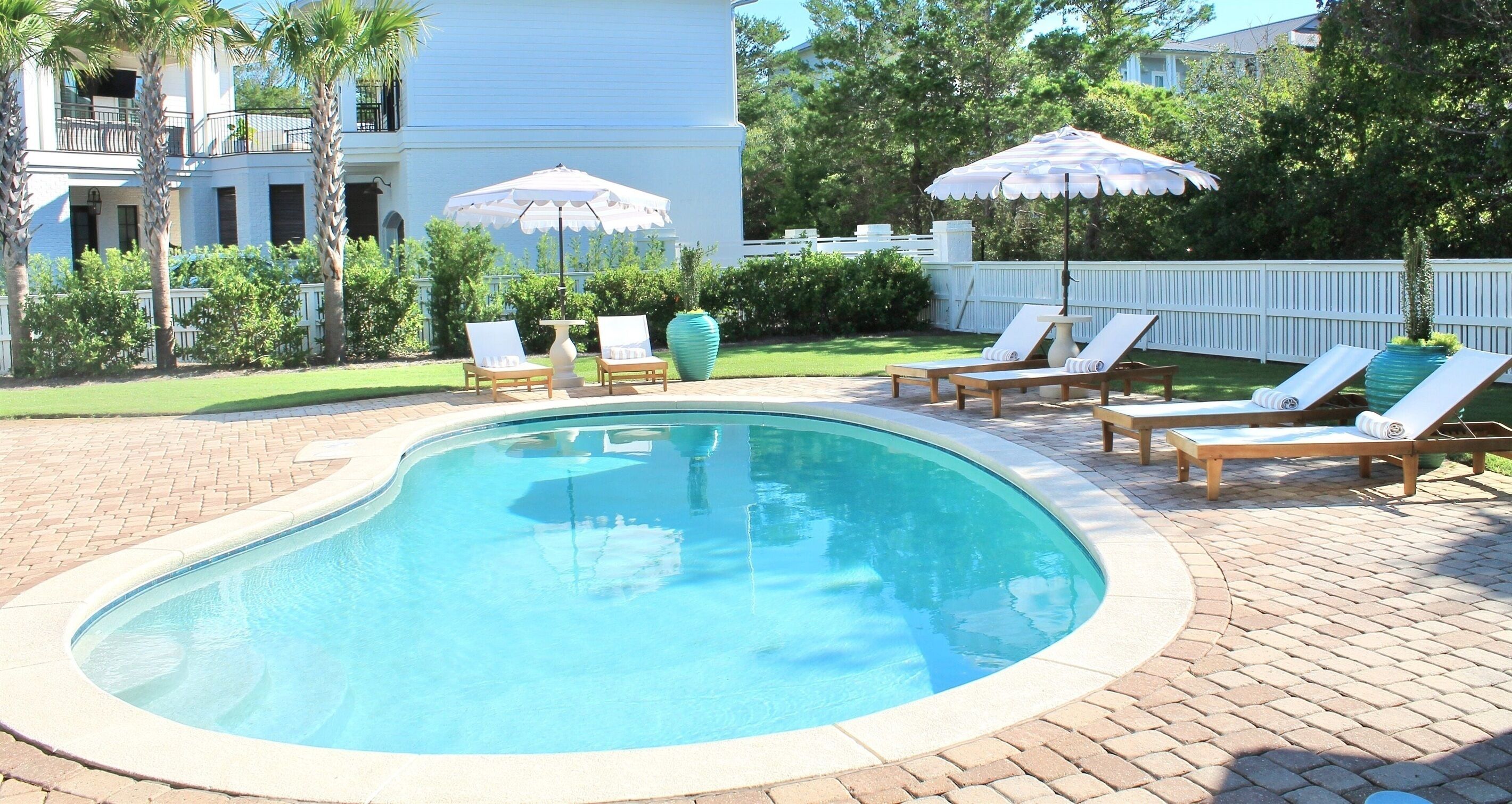 Une piscine entourée de chaises longues et de parasols, elle-même bordée d’une terrasse en briques et d’une clôture blanche.