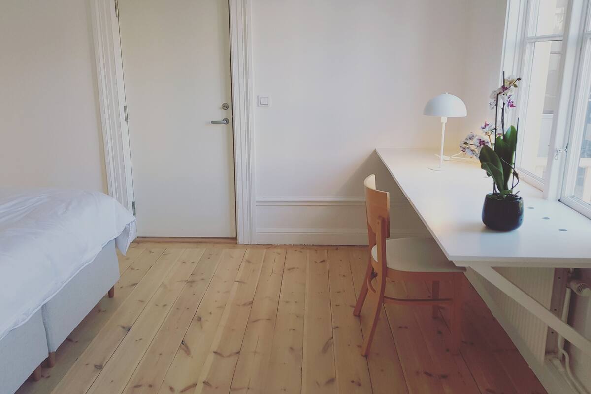 A bedroom with a wooden floor, a white bed, a desk with a chair, a potted orchid, and a window with a white lamp.