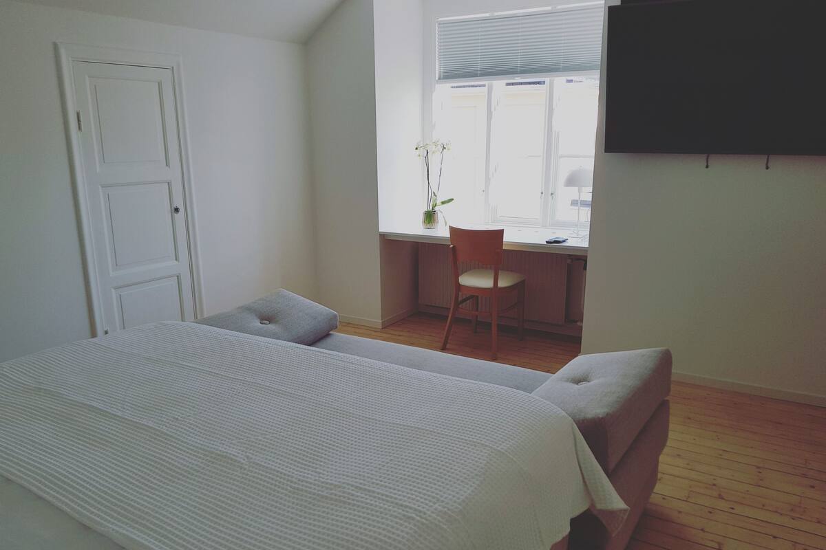 A bedroom with a bed, two pillows, a chair, a desk, and a window with blinds.