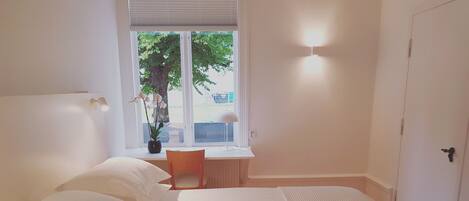 Quarto com cama, escrivaninha, cadeira e janela com vista para árvores e um edifício.