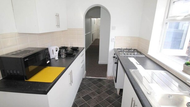 A compact kitchen with white cabinets, a black countertop, a microwave, a kettle, and a window providing natural light. - Townhouse @ Victoria Road Stoke (Stoke-on-Trent)