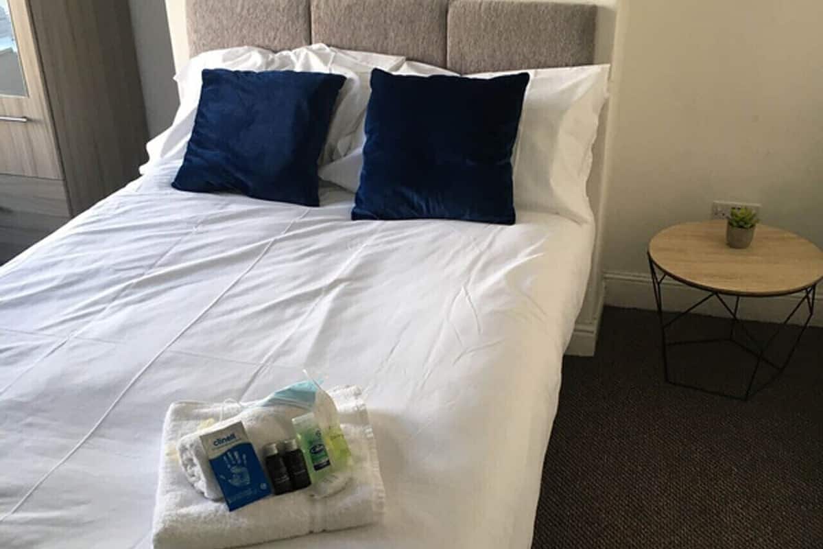 Una cama bien hecha con sábanas blancas y dos almohadas azul oscuro, una mesita redonda con una planta y un cuadro en la pared.