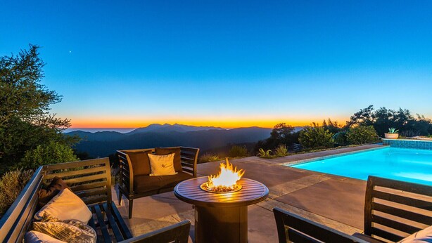 Zona de piscina con un hogar, mobiliario exterior y vistas a las montañas al atardecer.