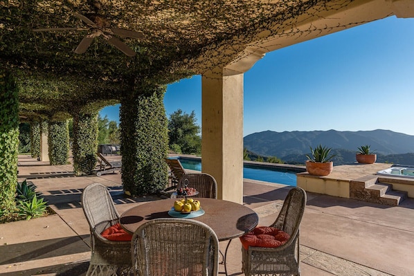 Um pátio externo com mobiliário de vime, uma mesa com frutas e vista para montanhas.