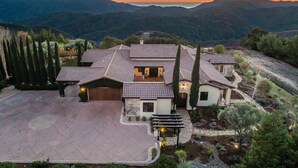 Una casa grande de varios pisos con techo de tejas, rodeada de montañas y árboles.
