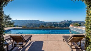 Piscina con vista a montañas, rodeada de sillones de descanso y una terraza con baldosas.