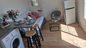 A compact living space with a washing machine, a dining table with chairs, a bed with pillows, a small table with a vase, and a refrigerator.