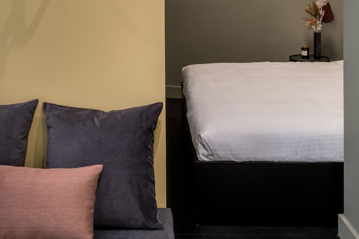 Un dormitorio con cama, sofá con cojines, lámpara de pared, una mesita con un jarrón y un arco de entrada.