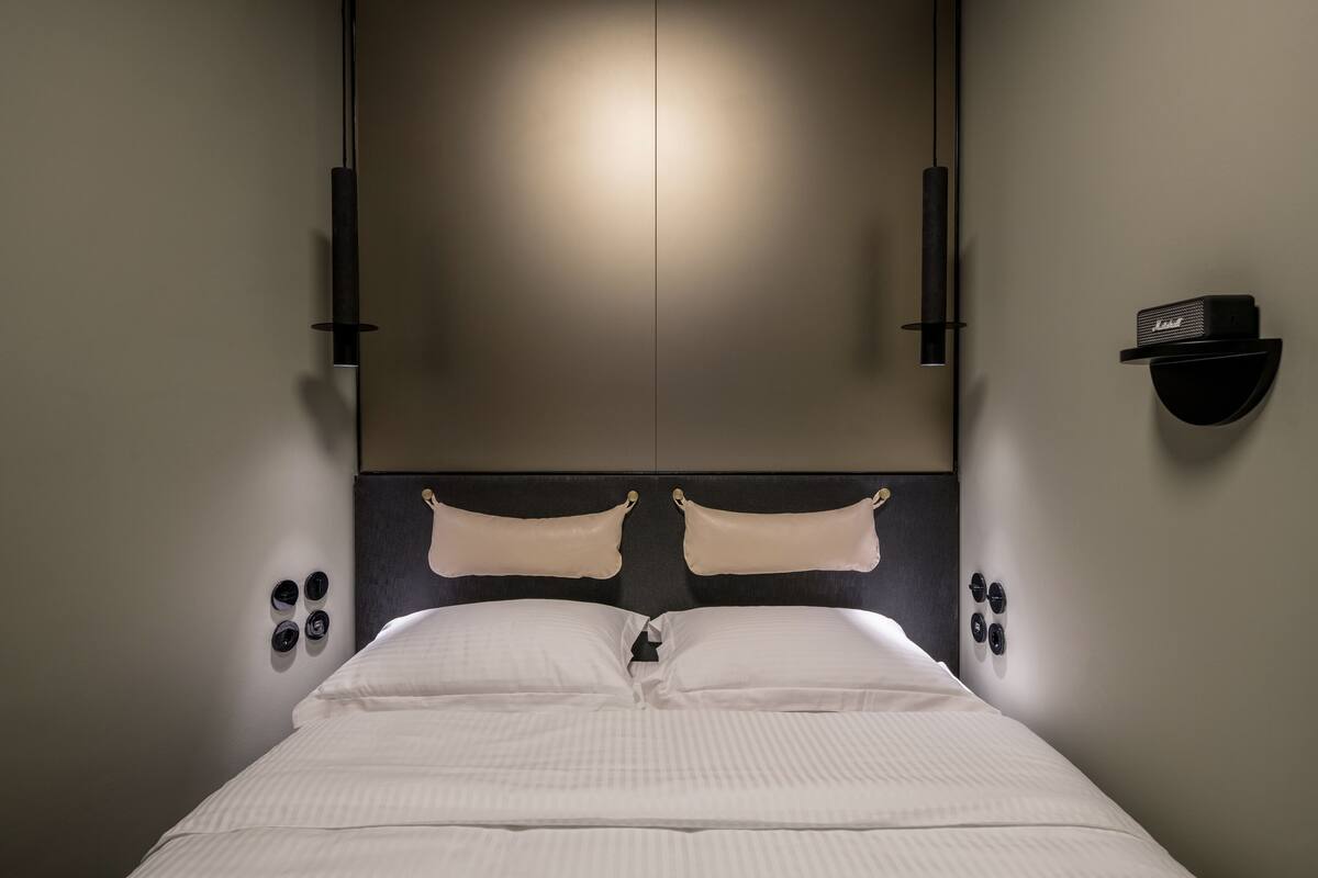 Una habitación de hotel moderna con una cama grande, cabecera con iluminación integrada, un altavoz montado en la pared y una pequeña estantería con un control remoto.