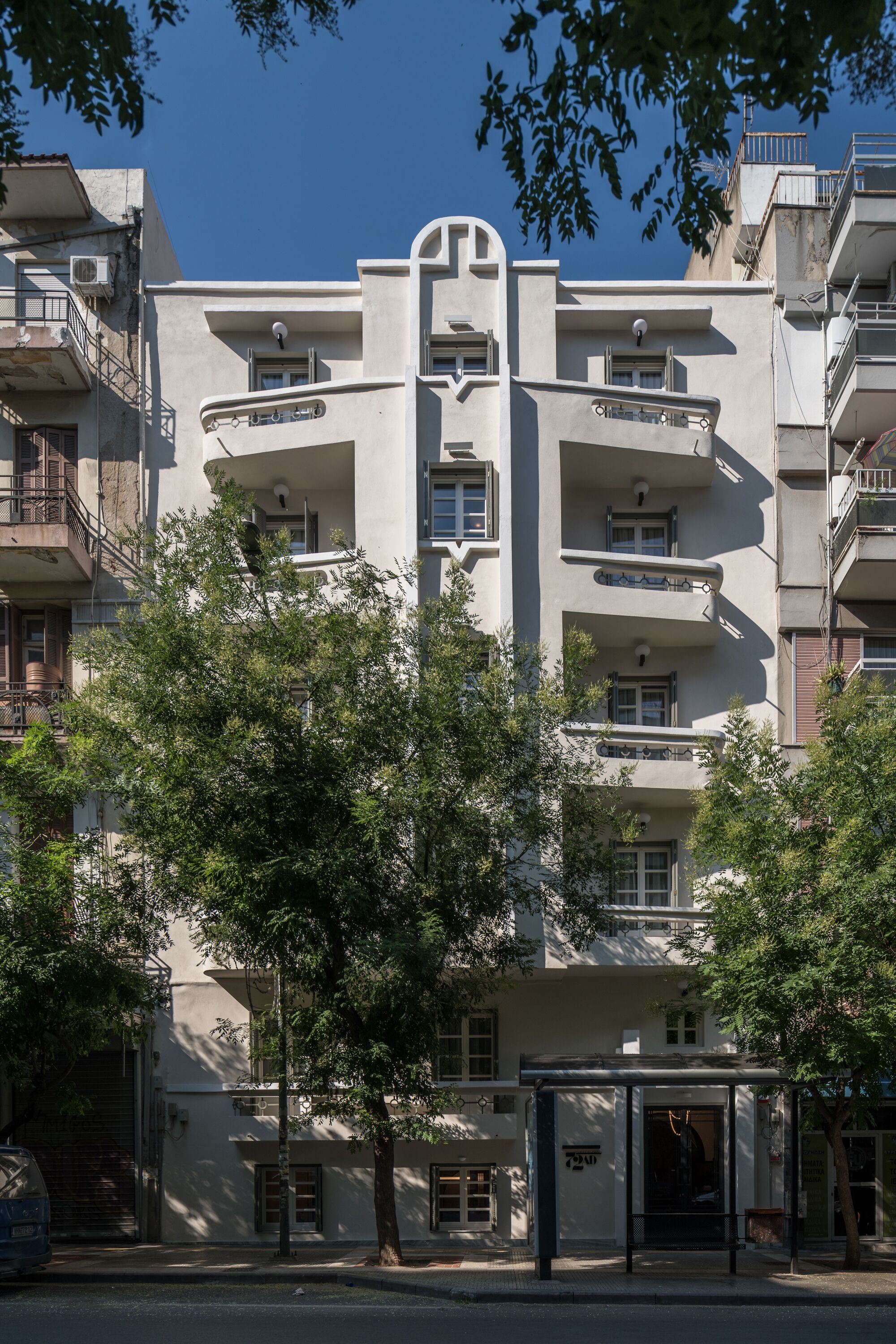 A multi-story building with balconies, a bus stop, and trees in front.