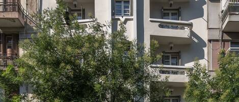 A multi-story building with balconies, a bus stop, and trees in front.
