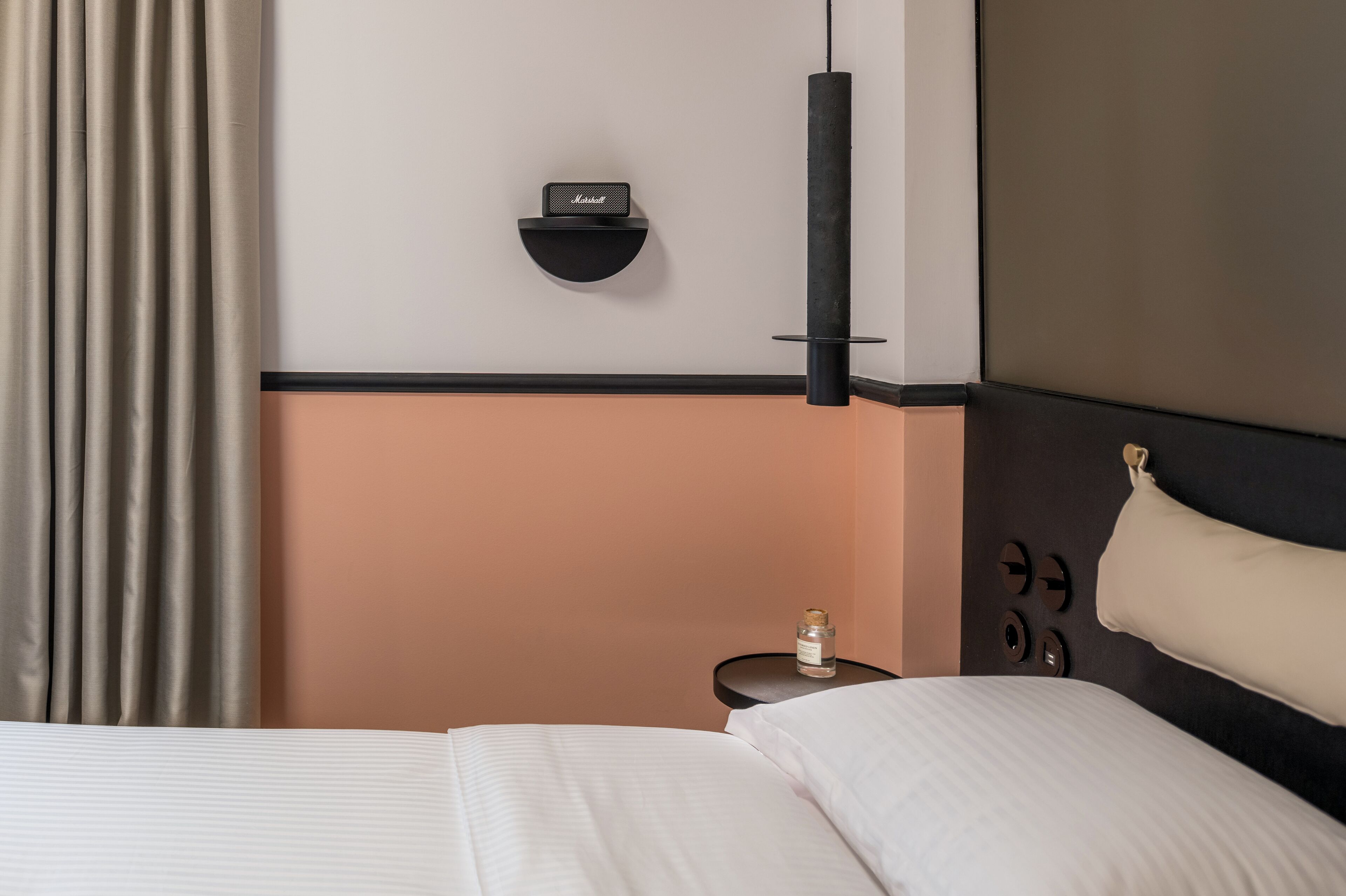 A hotel room with a bed, a nightstand, a wall-mounted speaker, and a hanging light fixture.