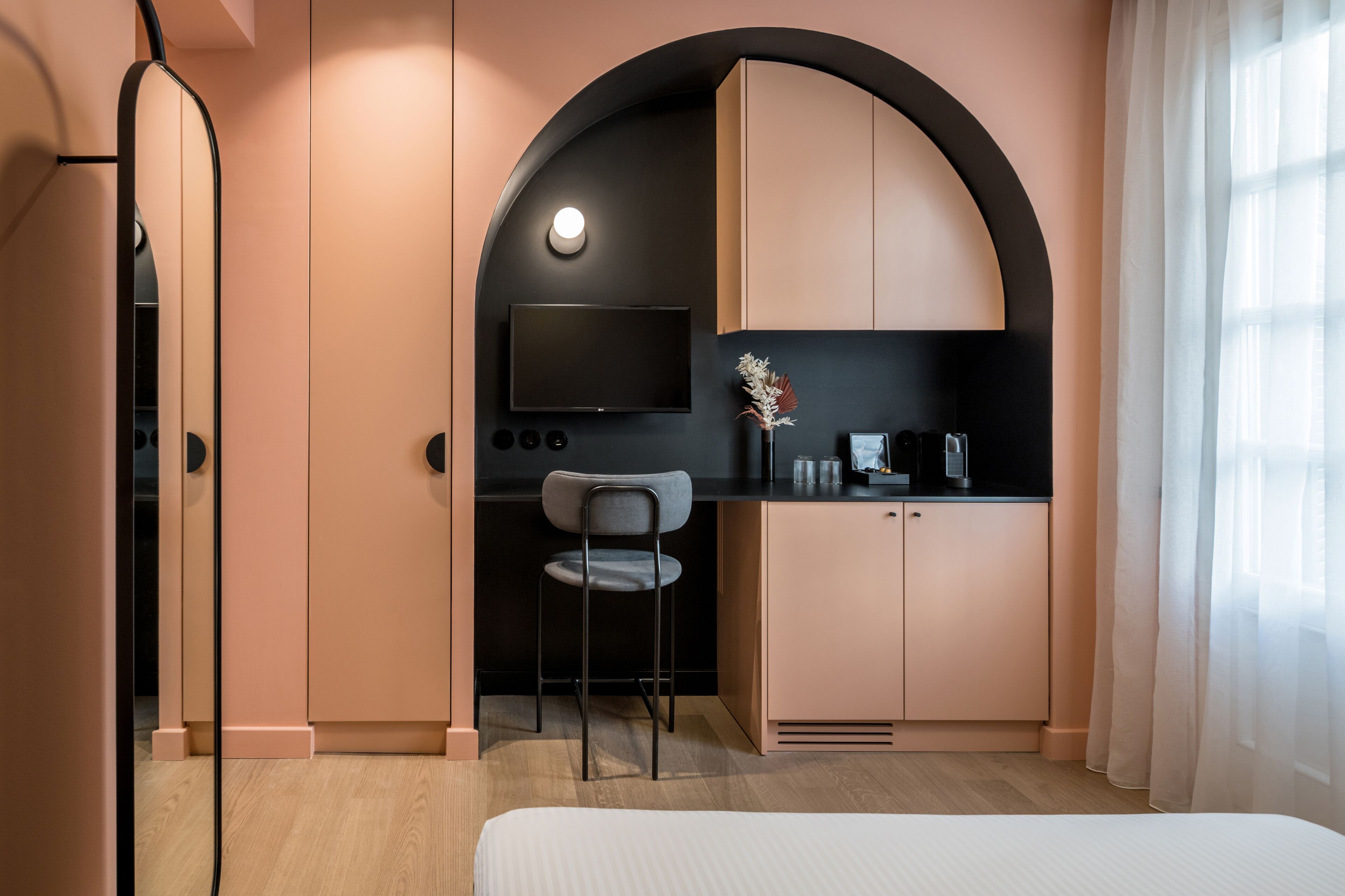 A modern hotel room with a bed, a desk with a chair, a TV, and a kitchenette.