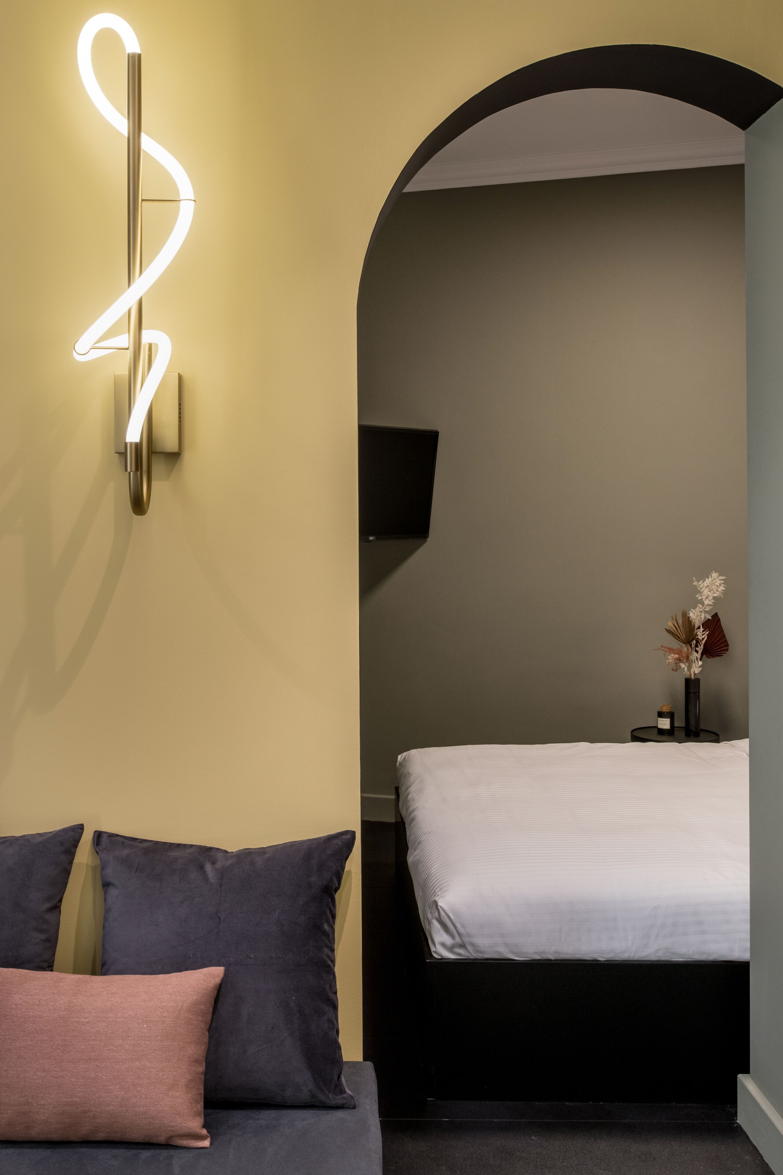 Un dormitorio moderno con una cama, un sofá, una lámpara de pared y una mesita con un jarrón.