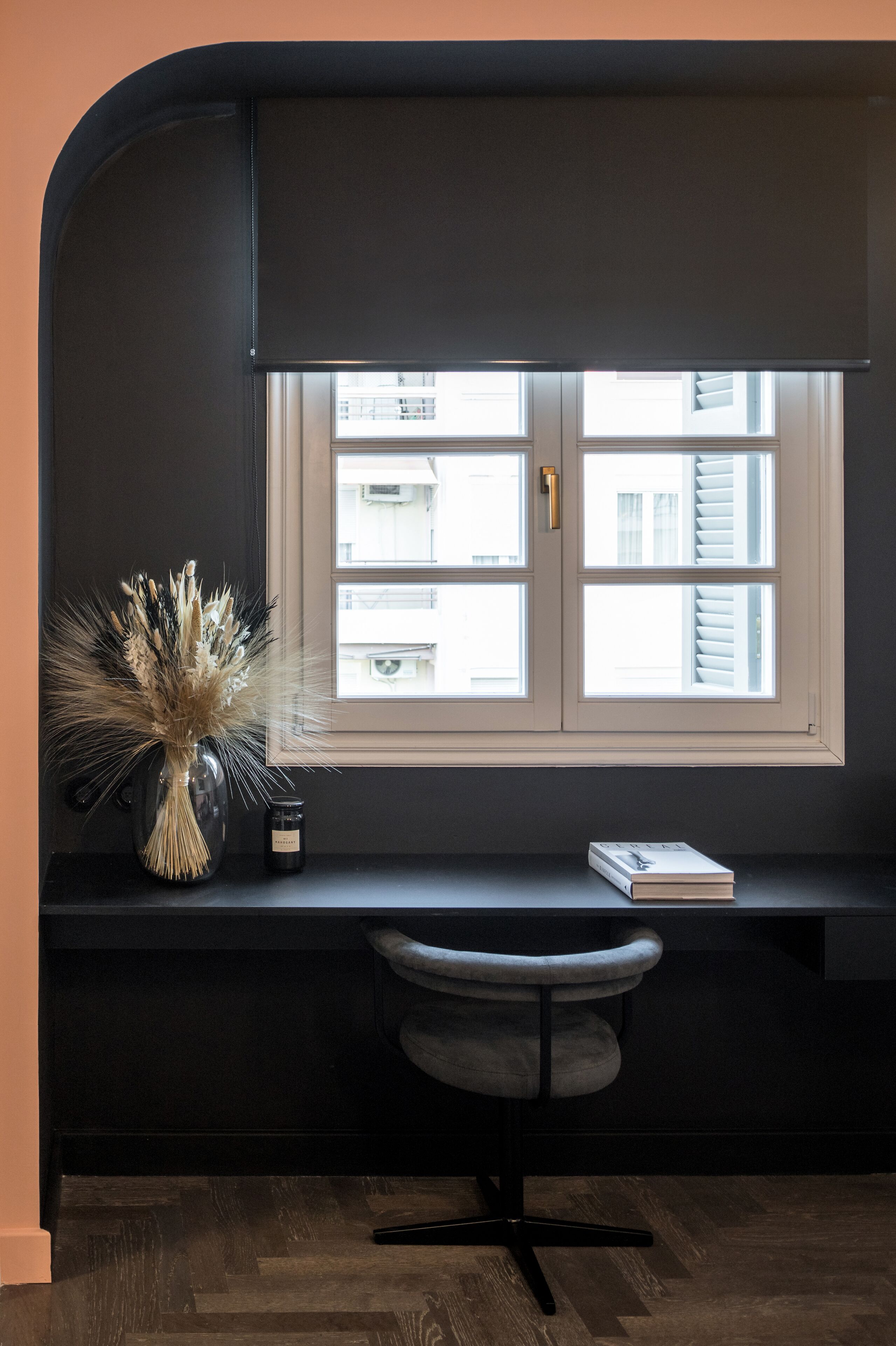 A room with a desk, a chair, a window with blinds, and a vase with dried plants.