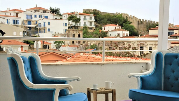 Un balcon avec deux chaises bleues et une petite table en bois, donnant sur une ville aux bâtiments aux toits de tuiles et sur une colline surmontée d’un fort.