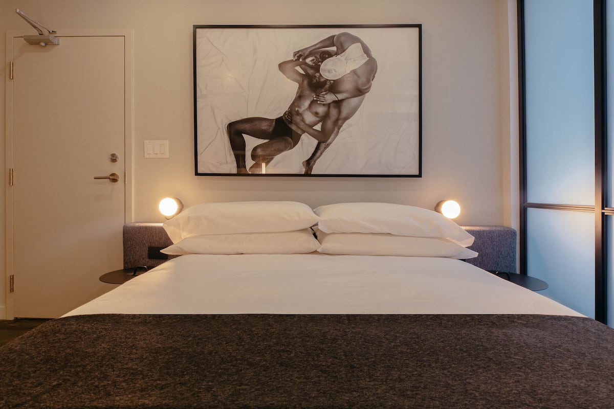 Cama com lençóis brancos e cabeceira marrom escura, quadro de uma figura musculosa na parede e luminárias de cabeceira.