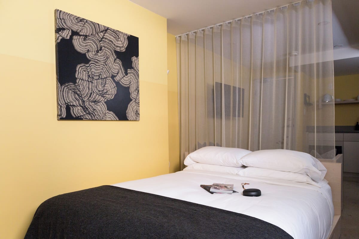 Quarto de hotel com uma cama, um quadro abstrato em preto e branco na parede e uma janela com cortinas leves.