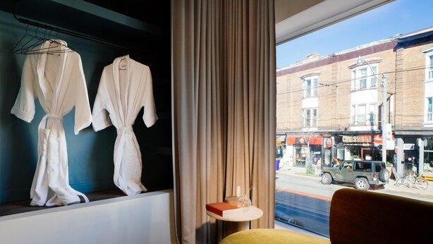 A hotel room with two white robes hanging on a rack, a small table with a candle, and a view of a street with a bus and buildings. - Ode (Toronto)