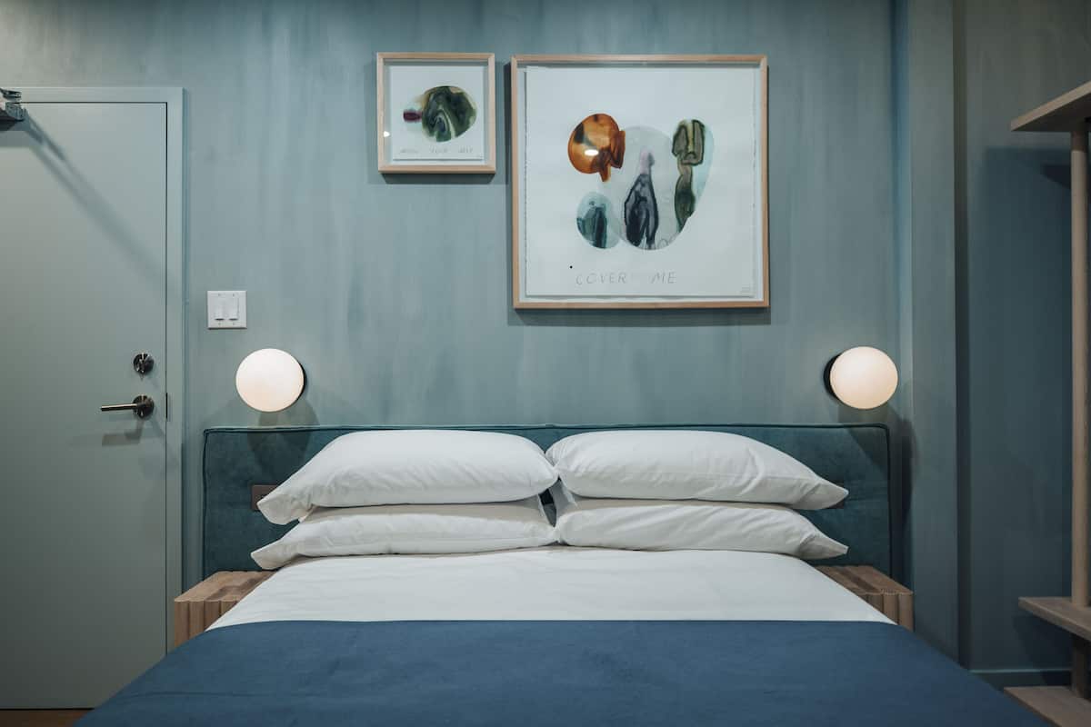 Cama bem arrumada com almofadas brancas e colcha azul em quarto com paredes azul-claras. Há duas obras de arte emolduradas acima da cama, uma com o texto "COVER ME" e outra com uma imagem menor.