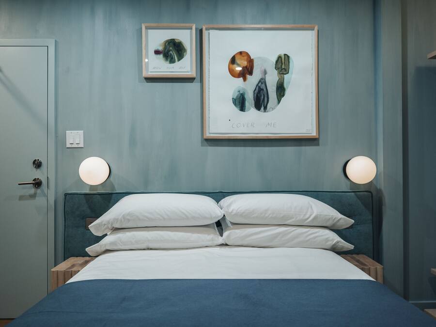 A neatly made bed with white pillows and a blue bedspread in a room with light blue walls. There are two framed artworks above the bed, one with a text that reads "COVER ME" and another with a smaller image.