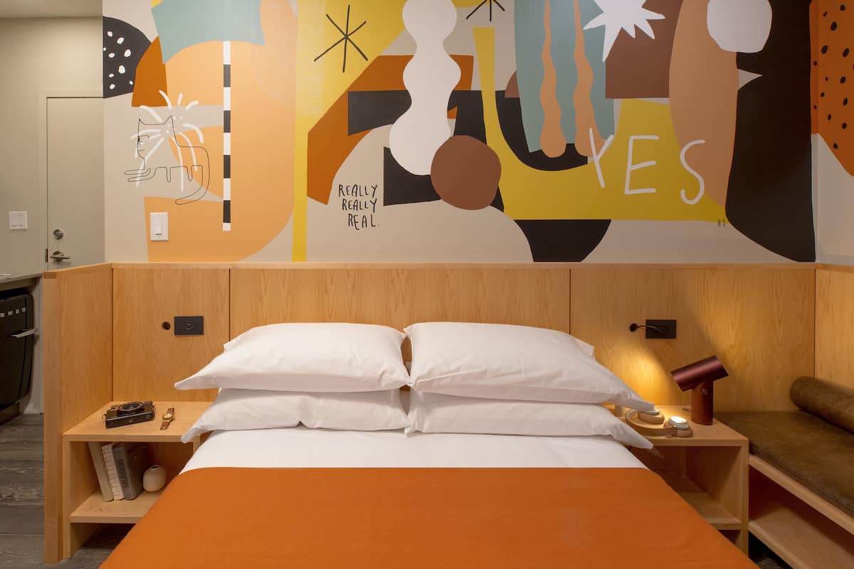 Quarto de hotel com cama, cabeceira de madeira, almofadas brancas, mesa de cabeceira com luminária, uma câmera e livros.