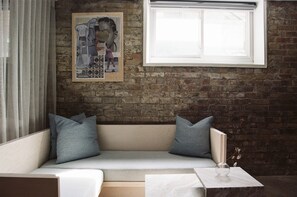 A cozy room with a brick wall, a white window, a beige sofa with blue cushions, and a small table with a glass vase.
