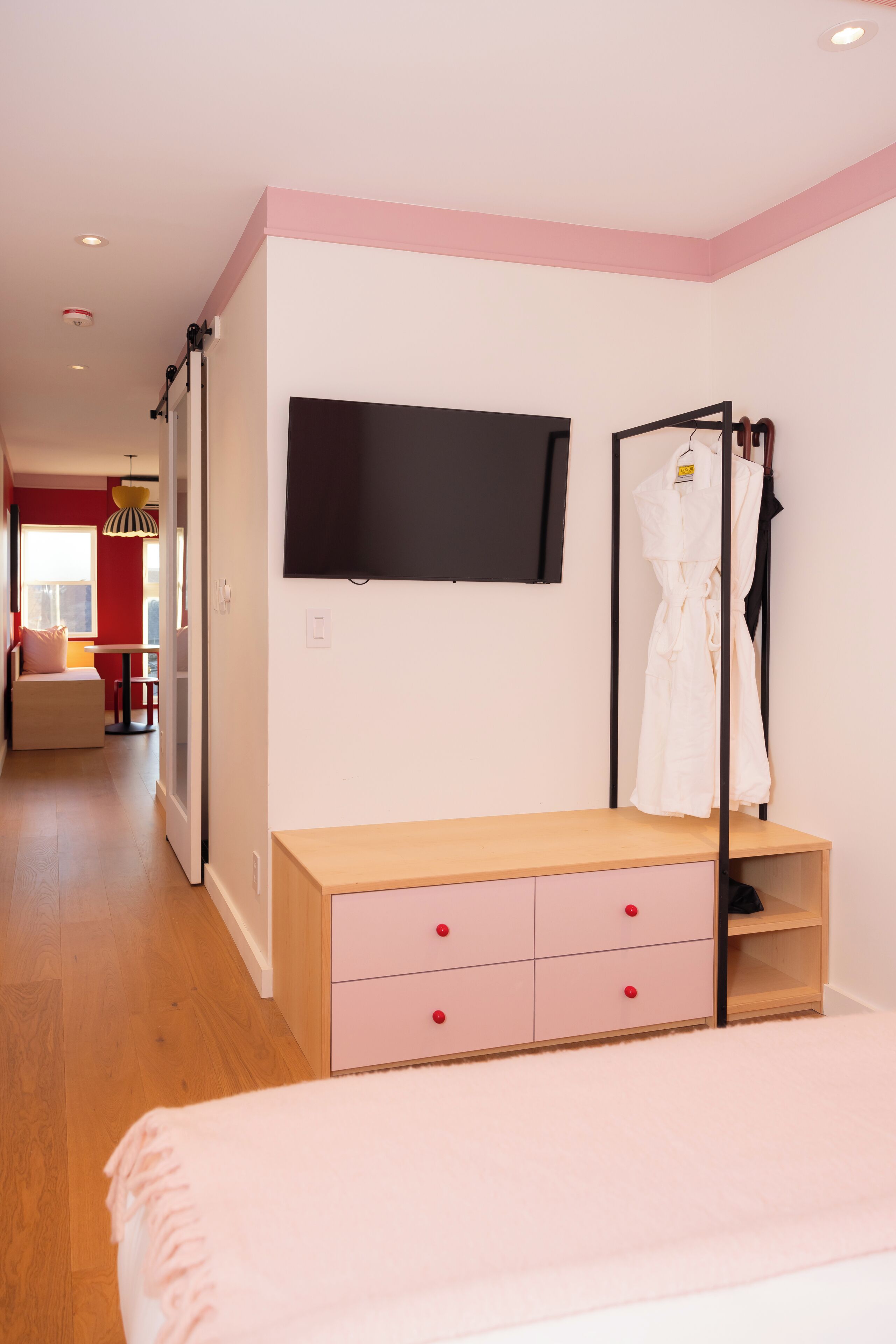 A bedroom with a bed, a TV mounted on the wall, a wooden dresser with drawers, and a robe hanging on a rack.