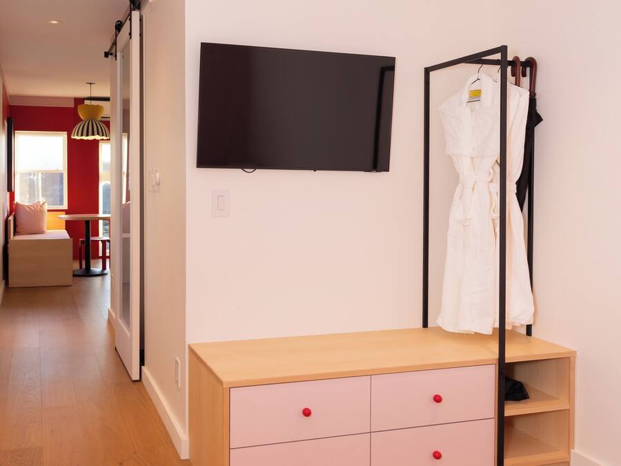 A bedroom with a bed, a TV mounted on the wall, a wooden dresser with drawers, and a robe hanging on a rack.